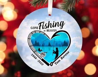 May include: A white round Christmas ornament with a blue heart design. The heart features a fishing scene with a silhouette of a person fishing. The text "Gone Fishing in Heaven" is written above the heart. The text "1948-2022 Tom Berens" is written below the heart.