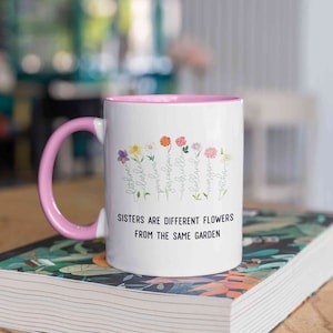 May include: White ceramic mug with a pink rim and handle. The mug features a floral design with the names of sisters and the text "Sisters are different flowers from the same garden."