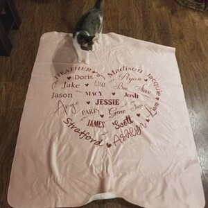 K&ouml;nnte beinhalten: Eine hellrosa Decke mit einem herzf&ouml;rmigen Design, das mit Namen in einer Schreibschrift gef&uuml;llt ist. Zu den Namen geh&ouml;ren Heather, Jake, Jason, Madison, Ryan und andere. Eine grau-wei&szlig;e Katze steht auf der Decke.