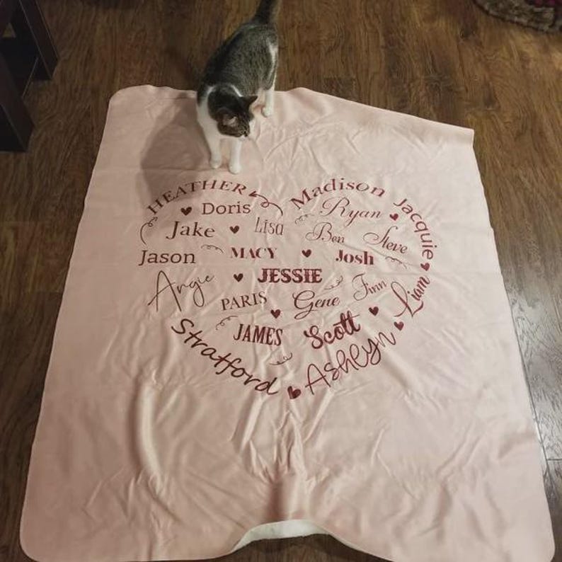 K&ouml;nnte beinhalten: Eine hellrosa Decke mit einem herzf&ouml;rmigen Design, das mit Namen in einer Schreibschrift gef&uuml;llt ist. Zu den Namen geh&ouml;ren Heather, Jake, Jason, Madison, Ryan und andere. Eine grau-wei&szlig;e Katze steht auf der Decke.