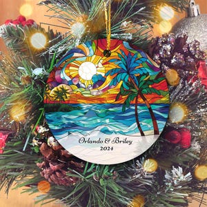 May include: Round ceramic Christmas ornament with a stained glass design of a beach scene with palm trees, a sunset, and ocean waves. The ornament is white with black text that reads "Orlando & Briley 2024".