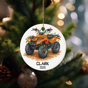 Peut inclure: Ornement de Noël rond avec un quad orange orné de guirlandes lumineuses colorées. L'ornement porte le nom "CLARK" et l'année "2025". Il est suspendu à une ficelle dorée.