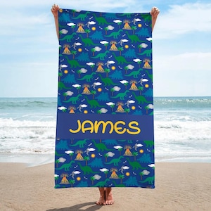 Peut inclure: Une serviette de plage bleue avec un motif répété de dinosaures verts, de volcans, de nuages et de soleils. La serviette a une bande bleu foncé avec le nom "JAMES" en lettres jaunes.