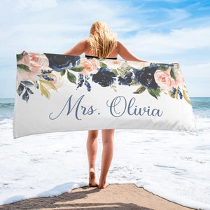 May include: A white beach towel with a floral design in shades of pink, blue, and green. The text "Mrs. Olivia" is printed in blue cursive script.