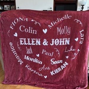 K&ouml;nnte beinhalten: Eine weinrote Fleece-Decke mit wei&szlig;em Text. Die Decke zeigt ein herzf&ouml;rmiges Design mit Namen wie "Ellen & John", "Fiona", "Colin" und anderen. Kleine wei&szlig;e Herzen sind ebenfalls im Design enthalten.