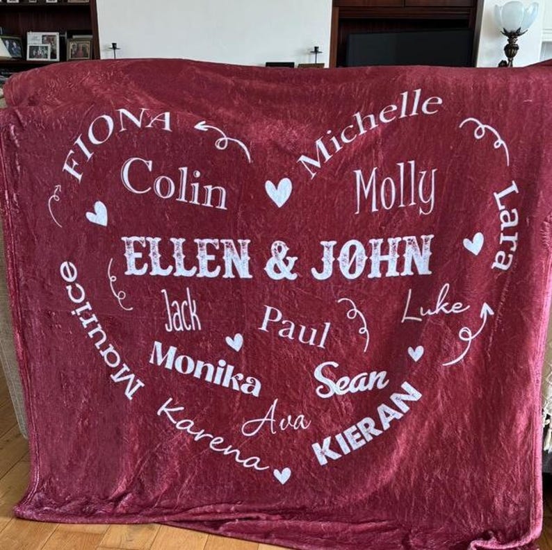 K&ouml;nnte beinhalten: Eine weinrote Fleece-Decke mit wei&szlig;em Text. Die Decke zeigt ein herzf&ouml;rmiges Design mit Namen wie "Ellen & John", "Fiona", "Colin" und anderen. Kleine wei&szlig;e Herzen sind ebenfalls im Design enthalten.