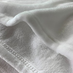 May include: Close-up of a white plush blanket with a stitched edge. The blanket is soft and fluffy, and the stitching is visible.