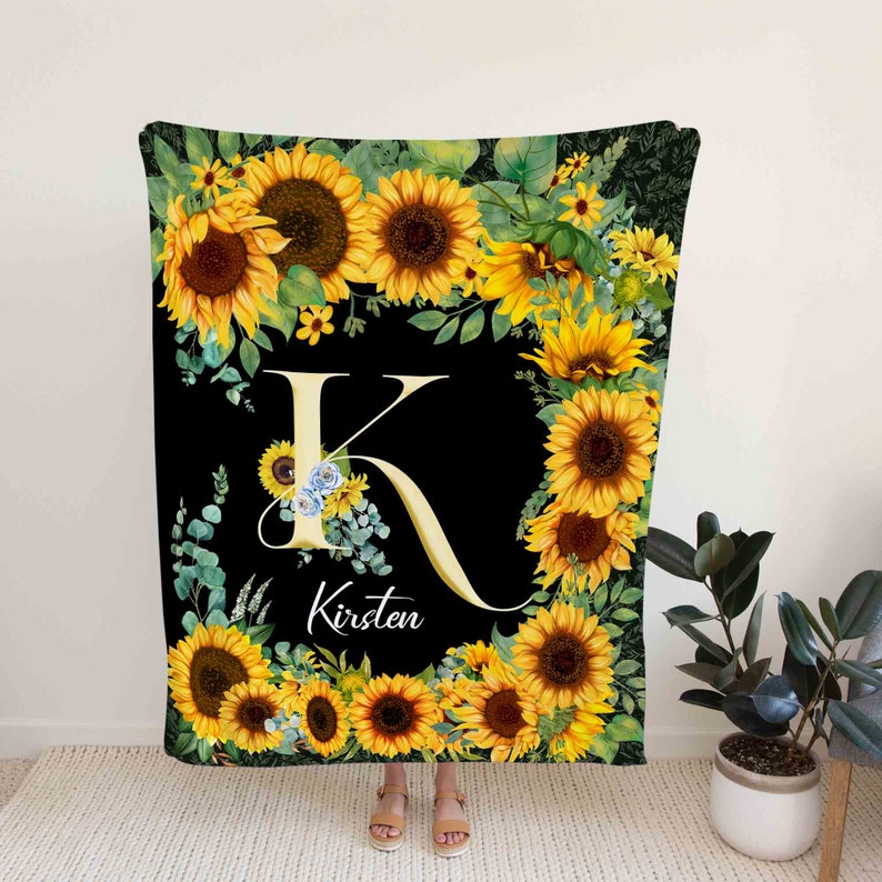 May include: A black throw blanket with a large yellow letter K surrounded by sunflowers and greenery. The name Kirsten is written below the letter K.