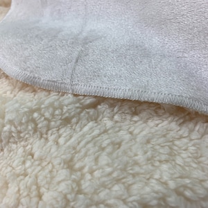 May include: Close-up of a white plush blanket with a soft, fluffy texture. The blanket has a white fleece lining on one side and a white sherpa lining on the other side.