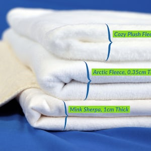 May include: A stack of three white fleece fabrics, each labeled with its type and thickness. The top layer is "Cozy Plush Fleece, 0.6cm Thick", the middle is "Arctic Fleece, 0.35cm Thick", and the bottom is "Mink Sherpa, 1cm Thick". A beige fabric is on the left.