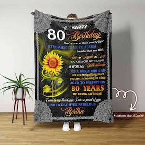 Puede incluir: Una manta negra y blanca con un diseño de girasol y el texto "Feliz 80 cumpleaños". El texto continúa con un mensaje sobre ser valiente, fuerte y una mujer con clase. La manta está personalizada con el nombre "Bertha" y el tamaño "Mediano (50x60)".