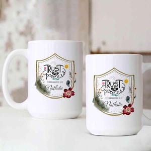 May include: Two white ceramic mugs with a decorative design. The design includes the words "Trust the Process" in script, a rainbow, and floral elements. The name "Nathalie" is also included. The mugs are different sizes.