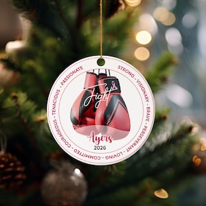 May include: A round Christmas ornament with a gold string, featuring red boxing gloves with black accents. The word "Fight" is in the center, with "Ayers 2026" below. The border includes words like "Courageous" and "Passionate".