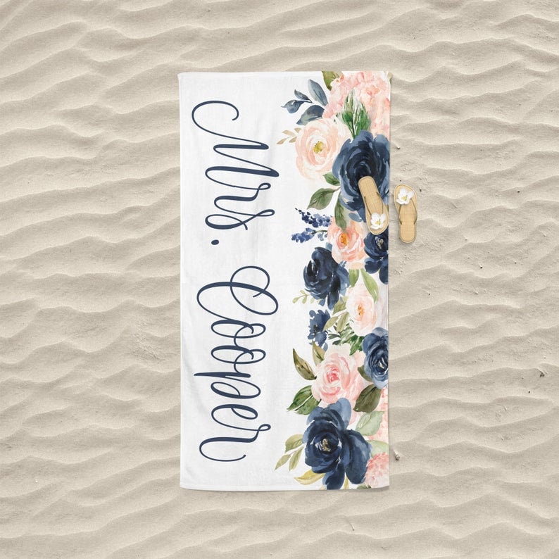 May include: White beach towel with a navy blue and pink floral design. The text "Mrs. Cooper" is written in navy blue cursive on the left side of the towel.
