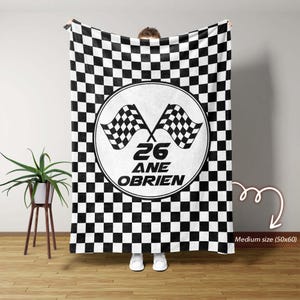 Könnte beinhalten: Eine schwarz-weiß karierte Decke mit einem kreisförmigen Design in der Mitte. Das Design zeigt zwei Zielflaggen, die Zahl 26 und den Namen "ANE OBRIEN". Die Decke ist mittelgroß, 127x152 cm.