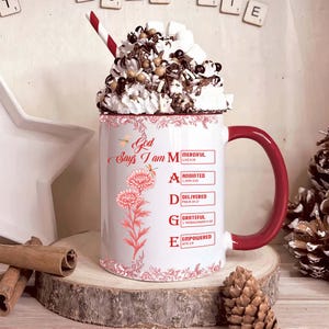 May include: A white ceramic mug with a red handle, filled with whipped cream, chocolate shavings, and marshmallows. The mug has a floral design and the text "God Says I am MADE" with related words. A red and white striped straw is in the drink.