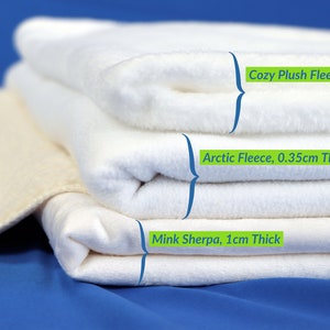 May include: Three white fleece blankets stacked on top of each other. The top blanket is labeled "Cozy Plush Fleece, 0.6cm Thick", the middle blanket is labeled "Arctic Fleece, 0.35cm Thick", and the bottom blanket is labeled "Mink Sherpa, 1cm Thick".