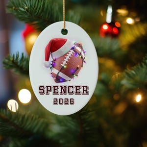 May include: Oval Christmas ornament featuring a football with a Santa hat and string lights. The ornament has the name "SPENCER" and the year "2026" printed in a collegiate font. The ornament hangs from a gold string.