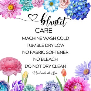 May include: A white card with floral borders and the text "blanket CARE" in black. The care instructions are listed below: "MACHINE WASH COLD, TUMBLE DRY LOW, NO FABRIC SOFTENER, NO BLEACH, DO NOT DRY CLEAN". The text "Hand made with love" is written in cursive at the bottom.