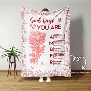 May include: White blanket with a red floral design and the words "God Says YOU ARE" in red. The blanket features a large red flower and the word "AMBER" with associated biblical verses. Butterflies and floral accents complete the design.
