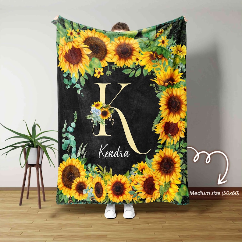May include: A black fleece blanket with a sunflower border and a large letter K in the center. The letter K is surrounded by a floral design and the name Kendra is written below it. The blanket is 50 inches by 60 inches.