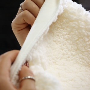 May include: A white sherpa fabric with a smooth white lining. The fabric is being held by a hand with red nail polish.