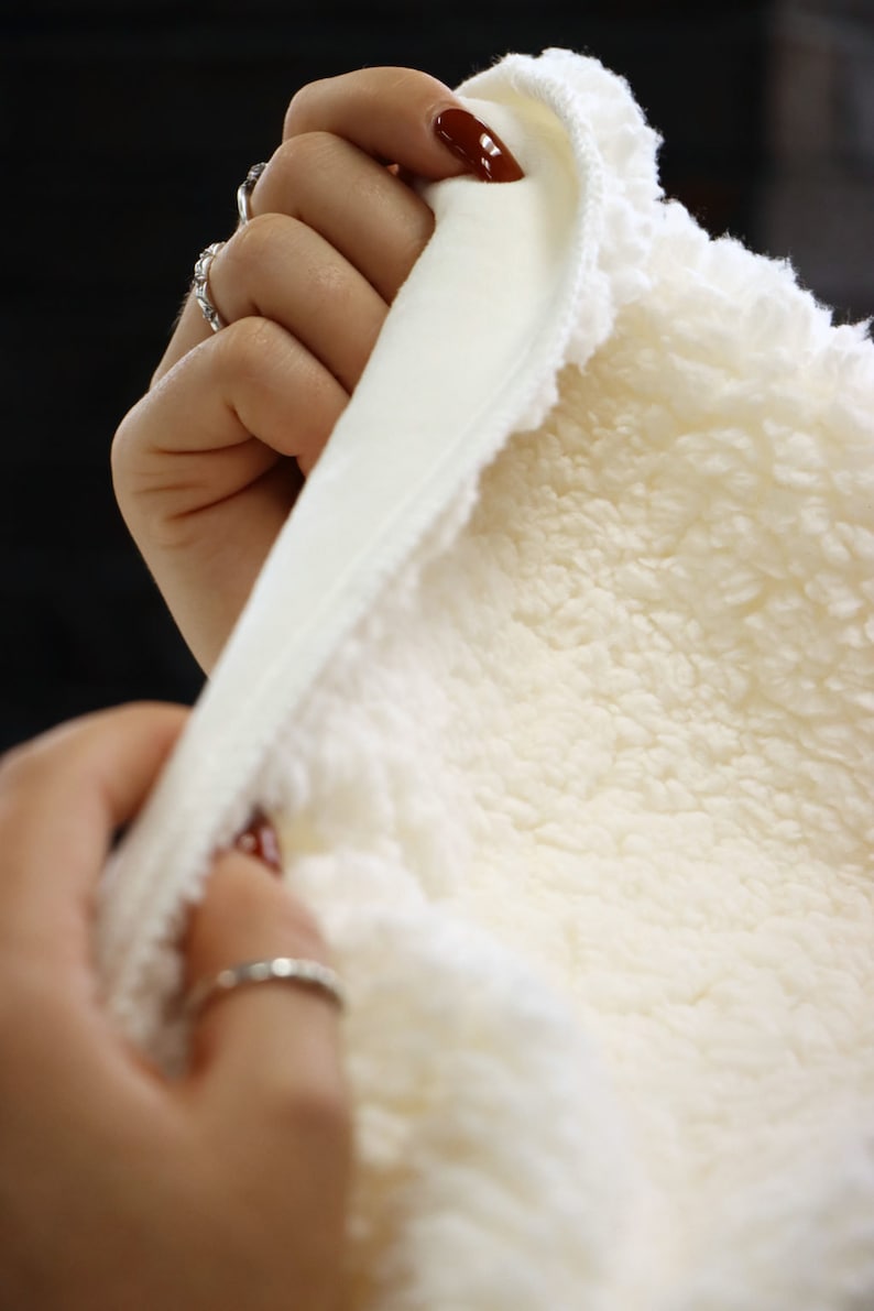 May include: A white sherpa fabric with a smooth white lining. The fabric is being held by a hand with red nail polish.