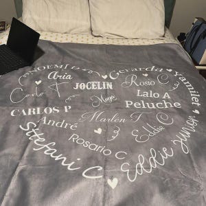 K&ouml;nnte beinhalten: Eine graue Decke mit einem herzf&ouml;rmigen Design, das mit Namen in wei&szlig;er Schrift gef&uuml;llt ist. Die Namen sind so angeordnet, dass sie das Herz bilden, mit kleinen Herzsymbolen. Die Decke liegt auf einem Bett.