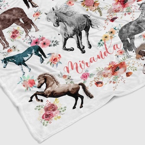 May include: A white fleece blanket with a watercolor design of horses and flowers. The name "Miranda" is written in pink script on the blanket.
