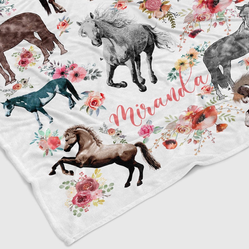 May include: A white fleece blanket with a watercolor design of horses and flowers. The name "Miranda" is written in pink script on the blanket.