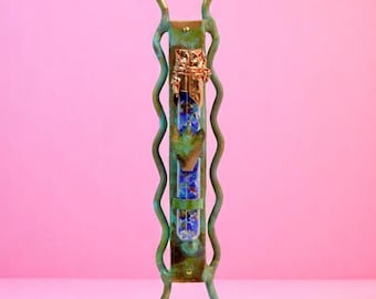 May include: A decorative mezuzah case with a green and gold finish. The case has a rectangular shape with wavy side supports and a gold-colored top. The center section features blue and gold accents. The mezuzah is standing on a pink background.