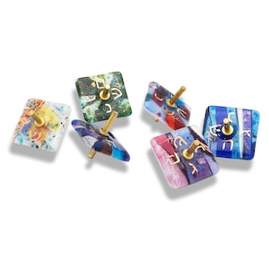 May include: A collection of colorful, square-shaped dreidels with gold-colored stems. The dreidels feature various designs and colors, including blue, green, red, and purple. Each dreidel has gold Hebrew letters on its surface.