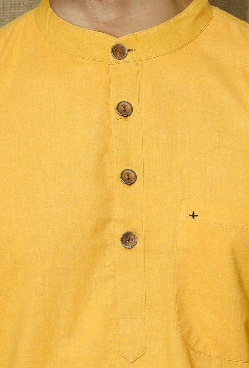 May include: A yellow linen shirt with a mandarin collar and wooden buttons. The shirt has a small pocket on the right side with a plus sign embroidered on it.