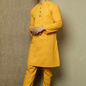 May include: A mustard yellow long-sleeved kurta with a button-up collar and a pocket on the left side. The kurta is paired with matching mustard yellow pants. The model is wearing brown leather sandals.