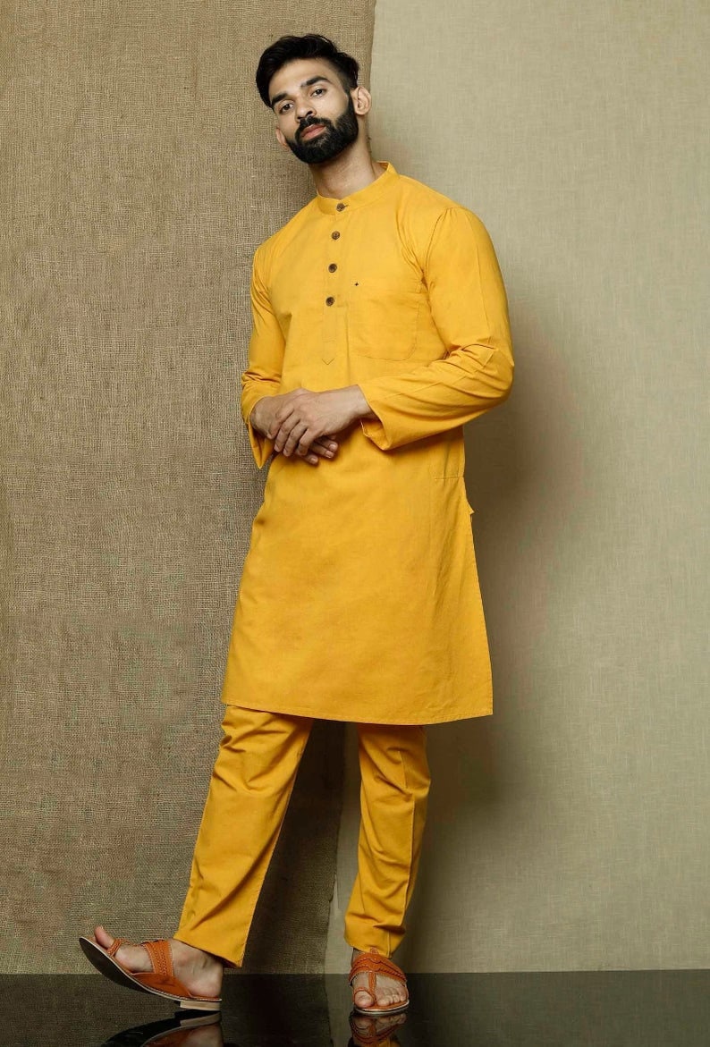May include: A mustard yellow long-sleeved kurta with a button-up collar and a pocket on the left side. The kurta is paired with matching mustard yellow pants. The model is wearing brown leather sandals.