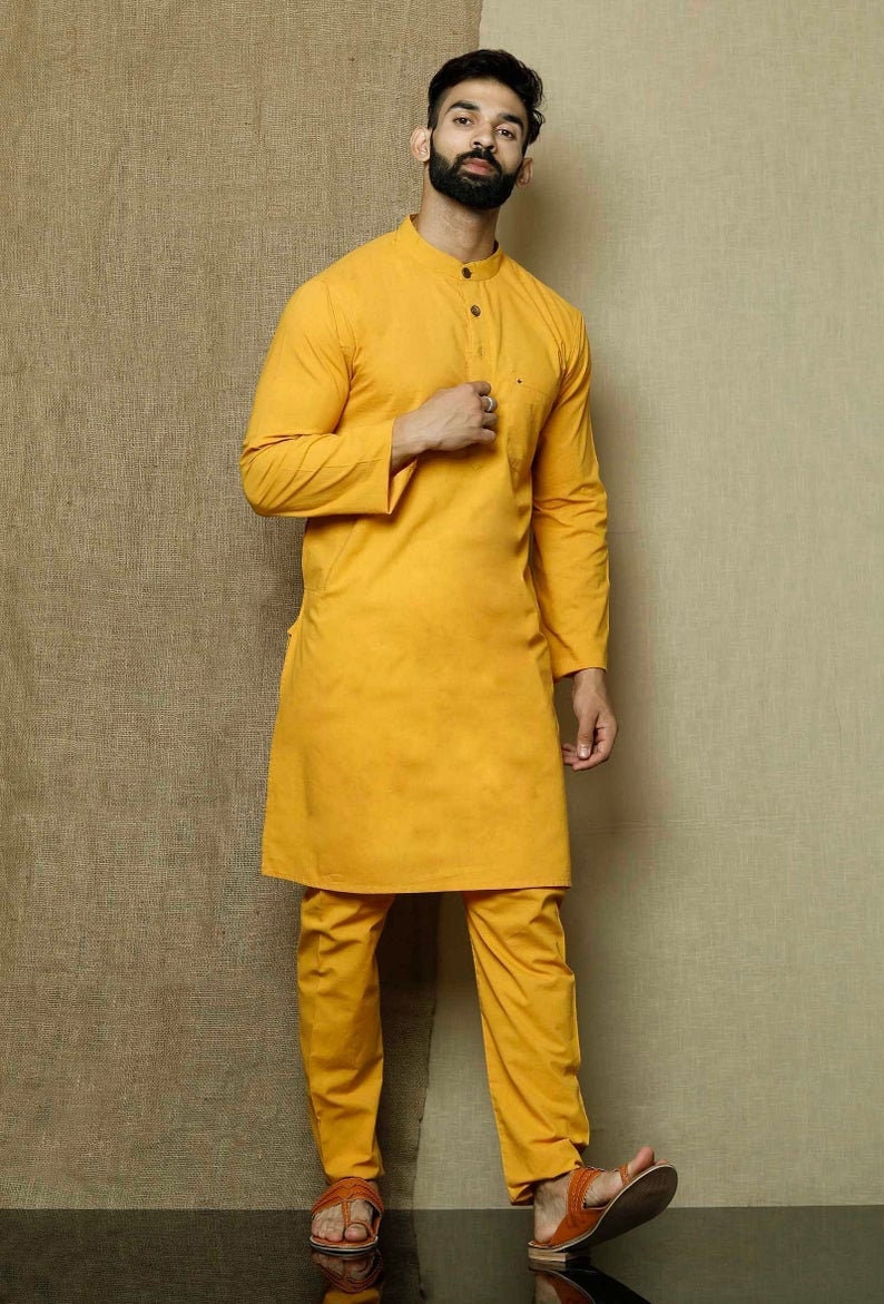 May include: A mustard yellow long-sleeved kurta with a mandarin collar and a button closure. The kurta is paired with matching mustard yellow pants. The model is wearing brown leather sandals.