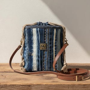 Könnte beinhalten: Eine Jeans-Beuteltasche mit braunem Lederriemen und Seildetails. Die Tasche hat ein Patchwork-Design mit blauem Denim, weißer Spitze und einem kleinen goldenen Etikett mit der Aufschrift "mi SHO". Die Tasche steht auf einer Holzoberfläche.