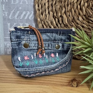Può includere: Un piccolo portamonete in denim con chiusura a zip e tiretto in pelle marrone. La borsa presenta fiori rosa ricamati e steli verdi. Il denim ha un lavaggio blu sbiadito.