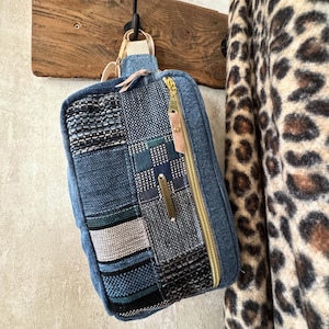 Może przedstawiać: Dżinsowa torba na ramię z patchworkowym wzorem w odcieniach niebieskiego, szarego i czarnego. Torba posiada złoty zamek błyskawiczny, beżowy pasek i małą skórzaną metkę. Torba wisi na drewnianym haczyku.