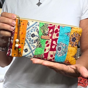 Może przedstawiać: Kolorowa kopertówka patchwork z złotym zamkiem błyskawicznym i skórzanym chwostem. Prostokątna torebka posiada pionowe panele z wzorzystej tkaniny w odcieniach czerwieni, błękitu, zieleni i żółci. Mała drewniana metka z napisem "hazelmade".
