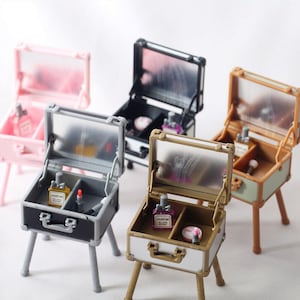 May include: Four miniature vanity cases with mirrors, each with a different color and design. The cases are open and show the interior compartments, which contain small bottles of perfume, lipstick, and other makeup items. The cases are on small wooden stands.