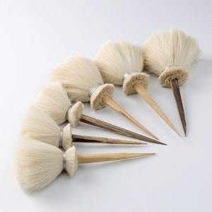 May include: Set of six small, white, round paint brushes with wooden handles. The brushes have a silver band around the base of the bristles.