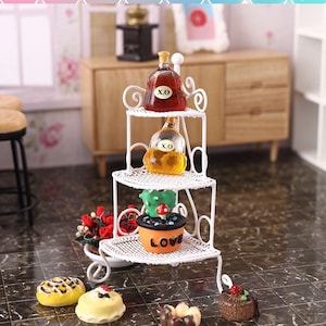 Może przedstawiać: Biała metalowa trójstopniowa półka narożna z dwiema szklanymi butelkami oznaczonymi "X.O." i doniczką z kaktusami z czerwonym grzybem na górze. Półka stoi na czarno-białej podłodze wyłożonej płytkami z kilkoma małymi ciastami.