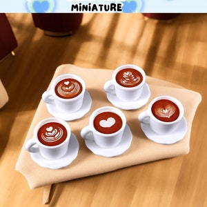 Può includere: Cinque tazzine da caffè bianche in miniatura con caffè marrone e disegni di latte art su una superficie beige. Le tazzine sono disposte in cerchio su una superficie beige. Il testo "STONE TOWER MINIATURE" è visibile nella parte superiore centrale dell'immagine.