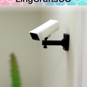 May include: A small white security camera with a black bracket fixed to a wall. The camera is rectangular with a dark lens. The bracket is black and attached to the wall. The text "LingCraftsCO" is at the top of the image.