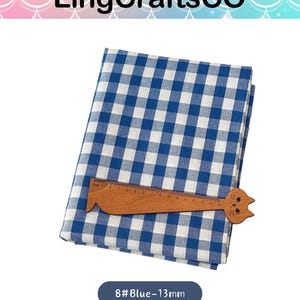 Puede incluir: Una pieza de tela doblada con un patr&oacute;n de cuadros vichy en azul y blanco. Una regla de madera con forma de gato est&aacute; colocada sobre la tela, mostrando una escala de medici&oacute;n de 0 a 15 cm. El texto 8#Blue-13mm es visible debajo de la tela.
