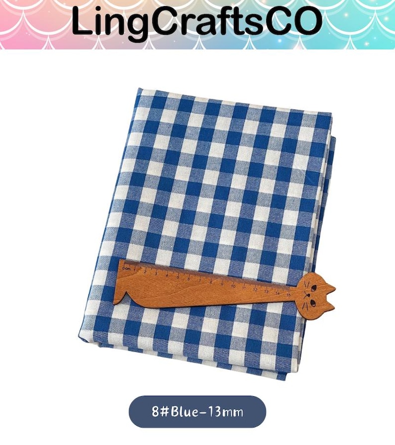 Puede incluir: Una pieza de tela doblada con un patr&oacute;n de cuadros vichy en azul y blanco. Una regla de madera con forma de gato est&aacute; colocada sobre la tela, mostrando una escala de medici&oacute;n de 0 a 15 cm. El texto 8#Blue-13mm es visible debajo de la tela.
