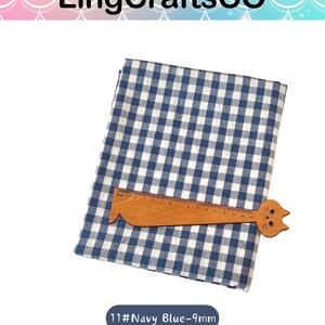 Puede incluir: Una pieza de tela doblada con un patr&oacute;n de cuadros vichy en azul marino y blanco. Una regla de madera con forma de gato est&aacute; colocada sobre la tela, con marcas claramente visibles hasta 15 cm. El texto 11#Navy Blue-9mm aparece debajo.