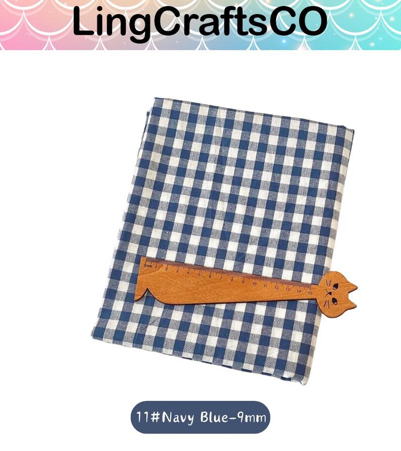Puede incluir: Una pieza de tela doblada con un patr&oacute;n de cuadros vichy en azul marino y blanco. Una regla de madera con forma de gato est&aacute; colocada sobre la tela, con marcas claramente visibles hasta 15 cm. El texto 11#Navy Blue-9mm aparece debajo.