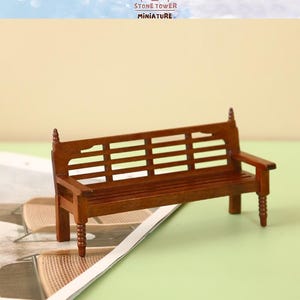 Peut inclure: Banc de parc miniature en bois brun foncé. Le banc présente un dossier et une assise à lattes, des accoudoirs et des détails décoratifs. Le texte "STONE TOWER MINIATURE" est visible. Le banc est posé sur un magazine.